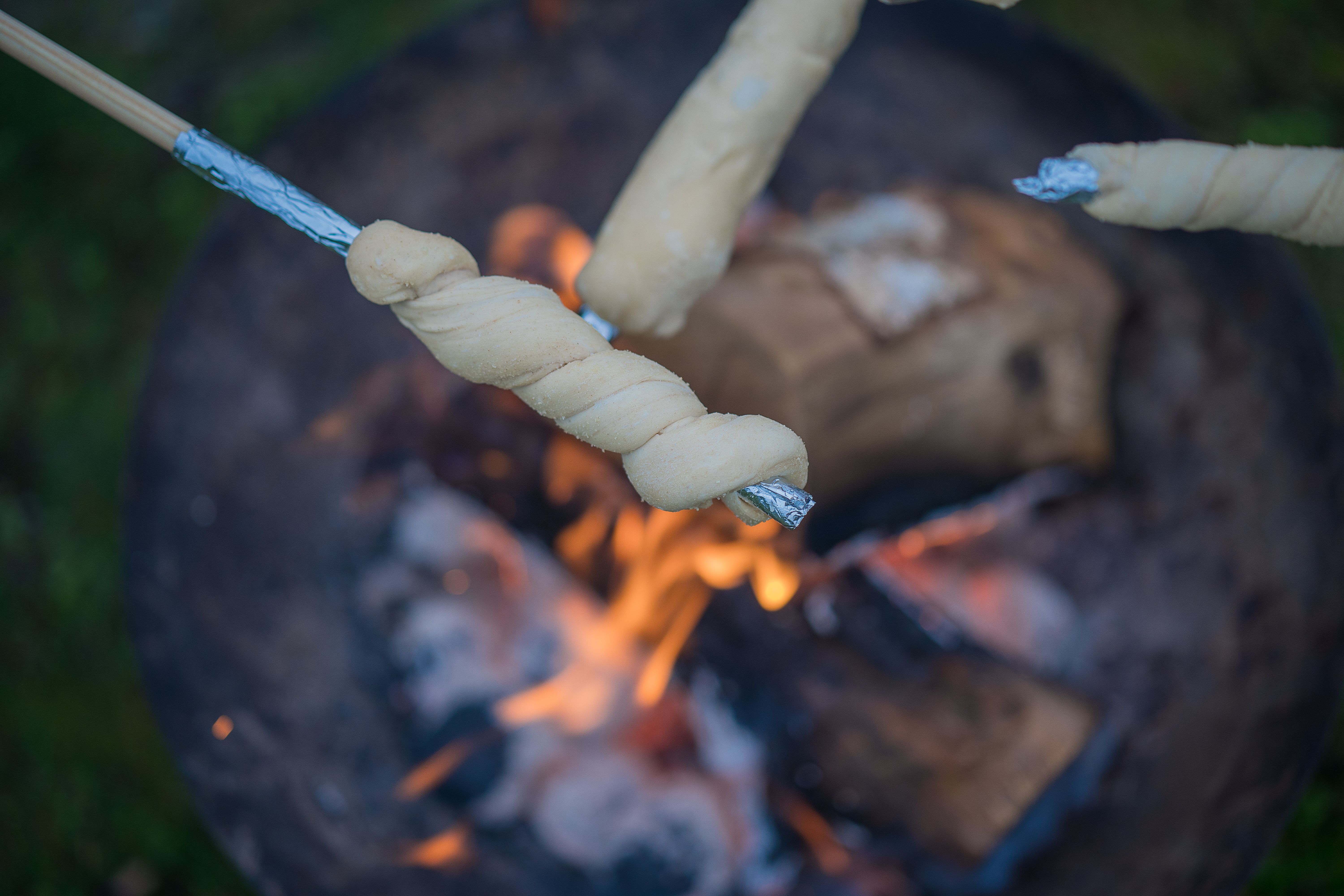Drei Stockbrote werden über einem offenen Lagerfeuer gebacken. Die Stöcke sind mit Alufolie umwickelt. Im Hintergrund sind Flammen und Glut zu sehen.