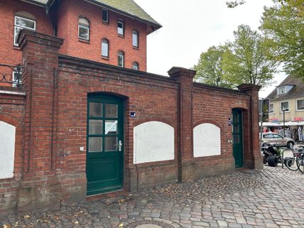 Backsteinmauer mit zwei grünen Türen und WC-Schildern, Kopfsteinpflaster davor. Im Hintergrund ein rotes Backsteingebäude und Bäume.