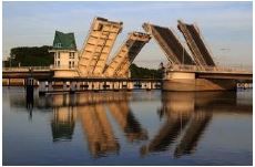 Geöffnete Klappbrücke spiegelt sich im ruhigen Wasser, ein Gebäude im Hintergrund. Die Szene zeigt eine friedliche Morgenstimmung.