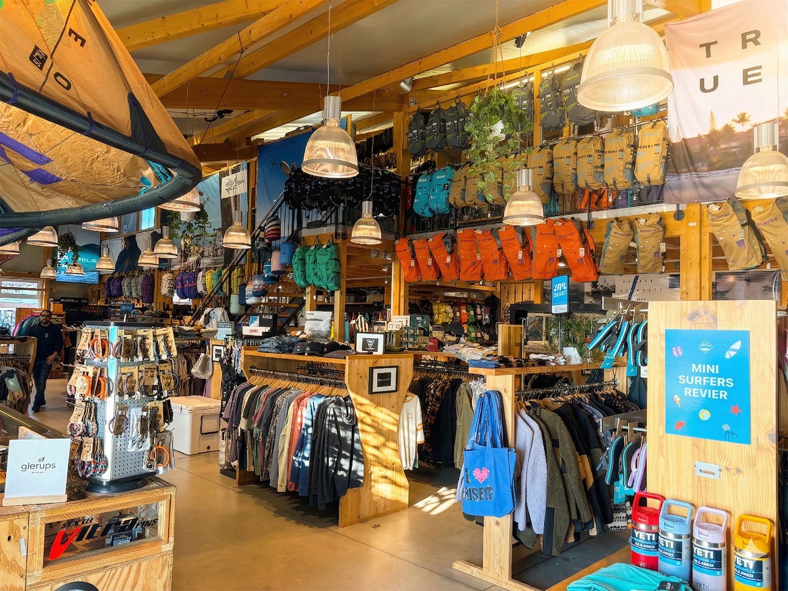 Ein gut sortierter Surfshop mit Kleidung, Ausrüstung und Accessoires. Bunte Taschen und Jacken hängen an den Wänden, Surfbretter sind dekorativ platziert.