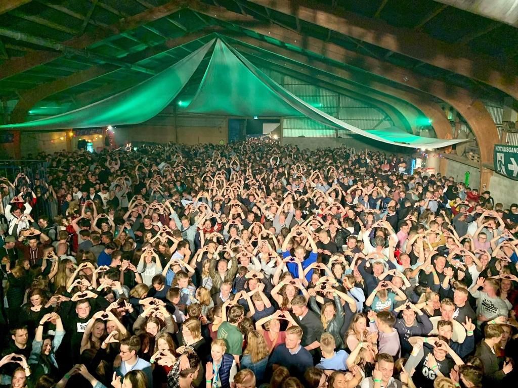 Eine große Menschenmenge in einer Halle formt mit den Händen Herzzeichen. Die Decke ist mit grünen Tüchern dekoriert.