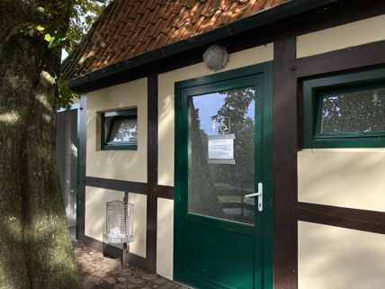 Eingang zu einer öffentlichen Toilette im Fachwerkstil mit grüner Tür und Behindertensymbol. Ein Mülleimer steht daneben, ein Baum wirft Schatten.