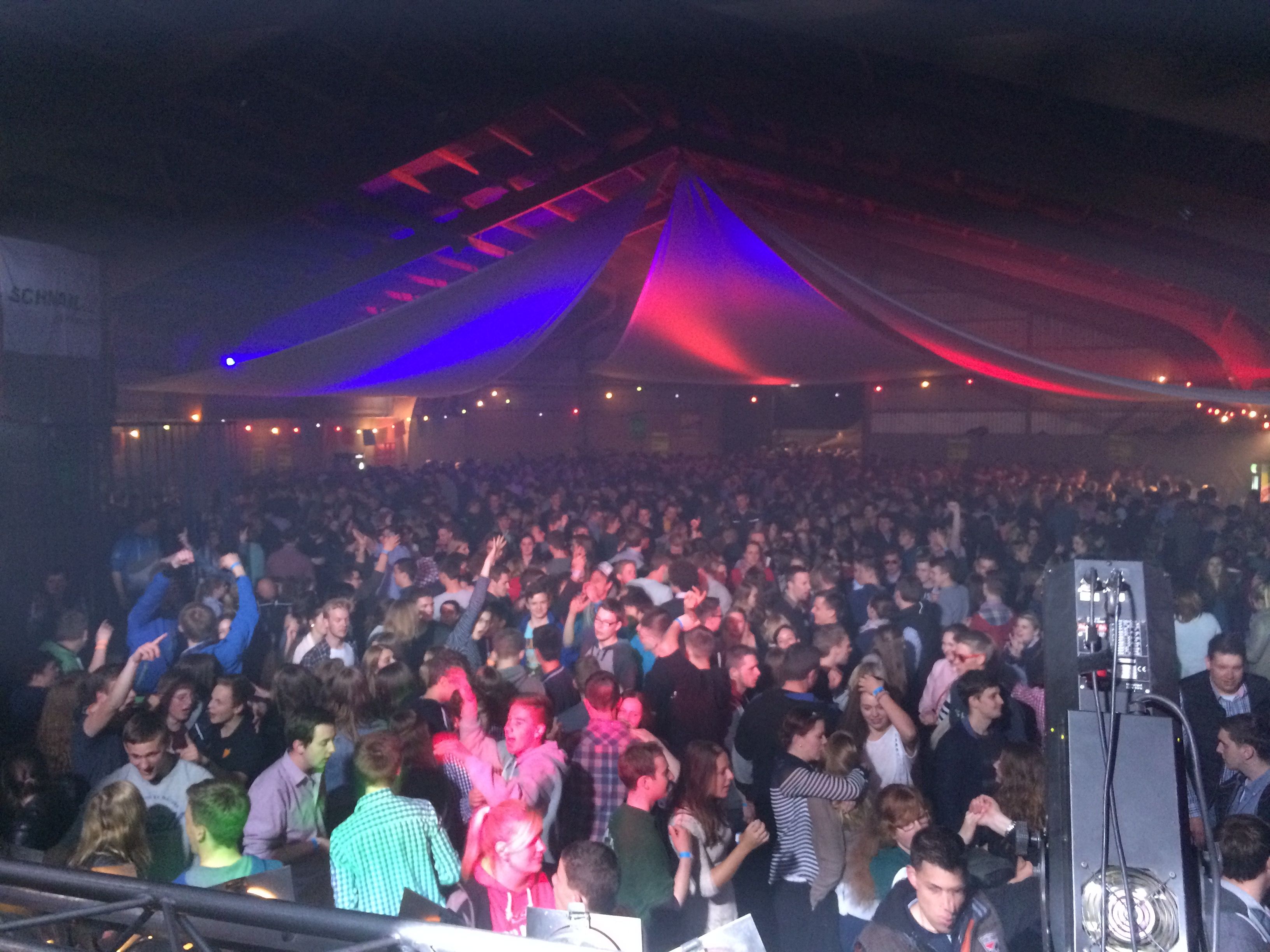 Eine große Menschenmenge tanzt in einer festlich beleuchteten Halle. Bunte Lichter und Dekorationen schaffen eine lebhafte Atmosphäre.