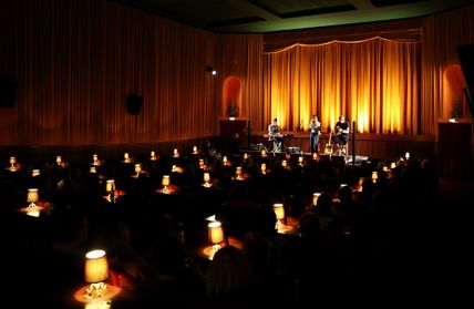 Ein Trio spielt auf einer Bühne in einem stimmungsvoll beleuchteten Kinosaal. Die Zuschauer sitzen an Tischen mit kleinen Lampen.
