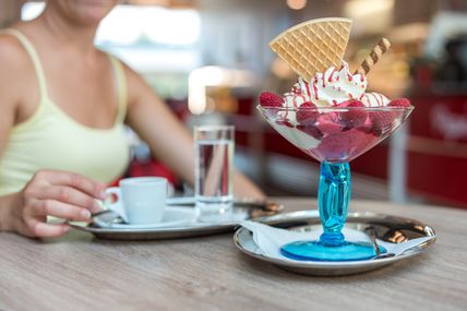 Ein köstliches Dessert mit Eiscreme, Himbeeren und Waffeln in einem blauen Glas. Im Hintergrund genießt eine Person eine Tasse Kaffee.