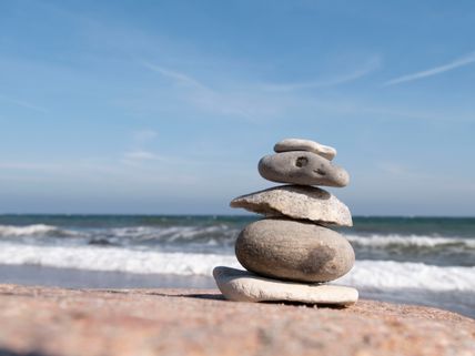 Ein Steinmännchen aus flachen Steinen steht am Strand vor einem welligen Meer und blauem Himmel. Es vermittelt Ruhe und Balance.