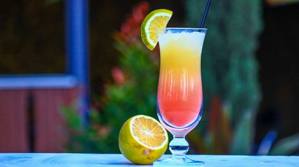Ein Cocktailglas mit einem bunten Getränk und Zitronenscheibe, daneben eine halbe Zitrone auf einem Tisch. Der Hintergrund ist unscharf.