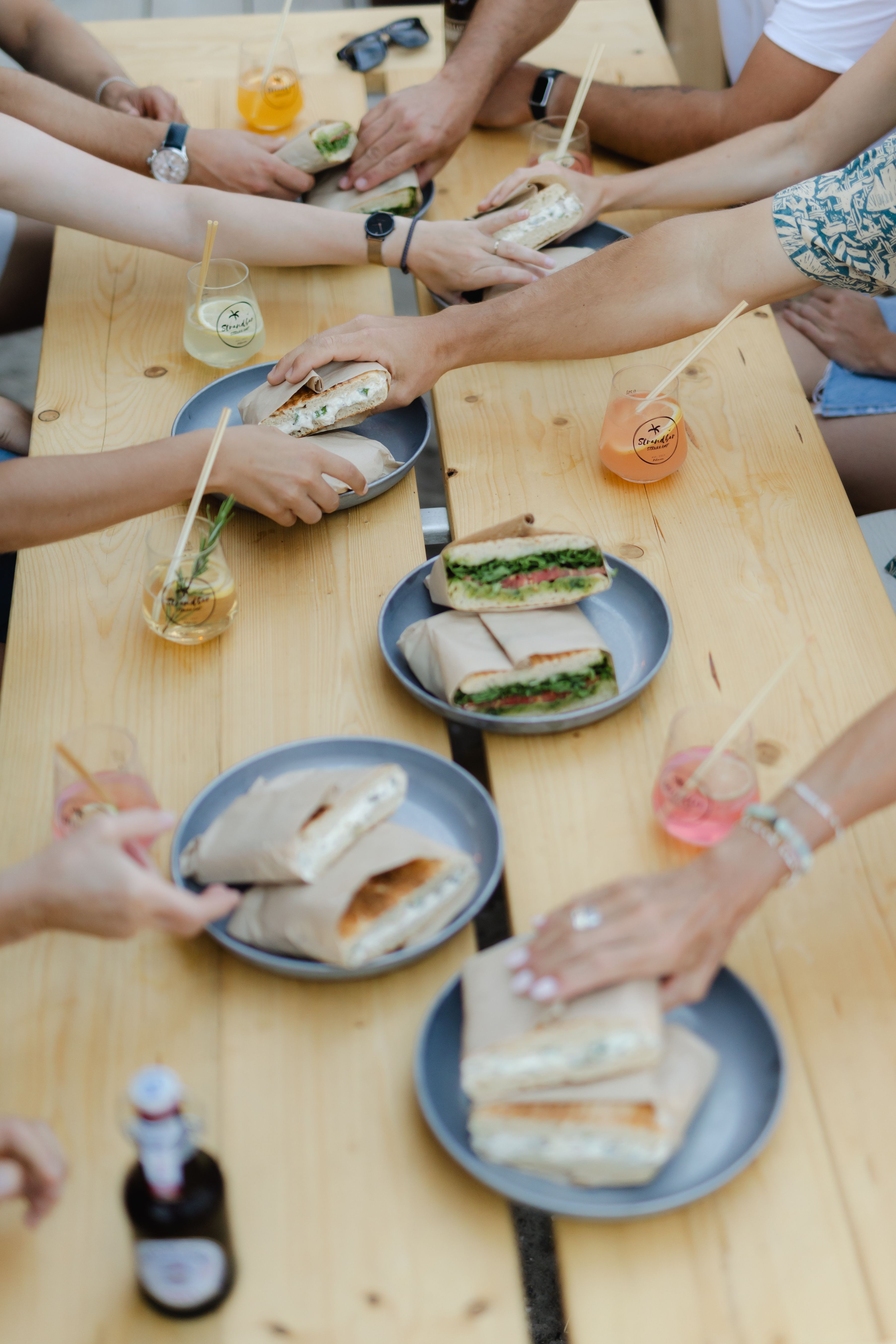 Freunde teilen Sandwiches und Getränke an einem Holztisch. Verschiedene Getränke in Gläsern mit Strohhalmen sind zu sehen.