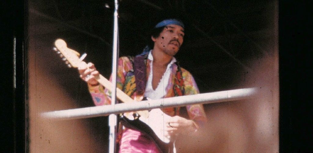 Ein Musiker mit buntem Hemd und Stirnband spielt E-Gitarre auf einer Bühne. Im Hintergrund ist ein Mikrofonständer zu sehen.