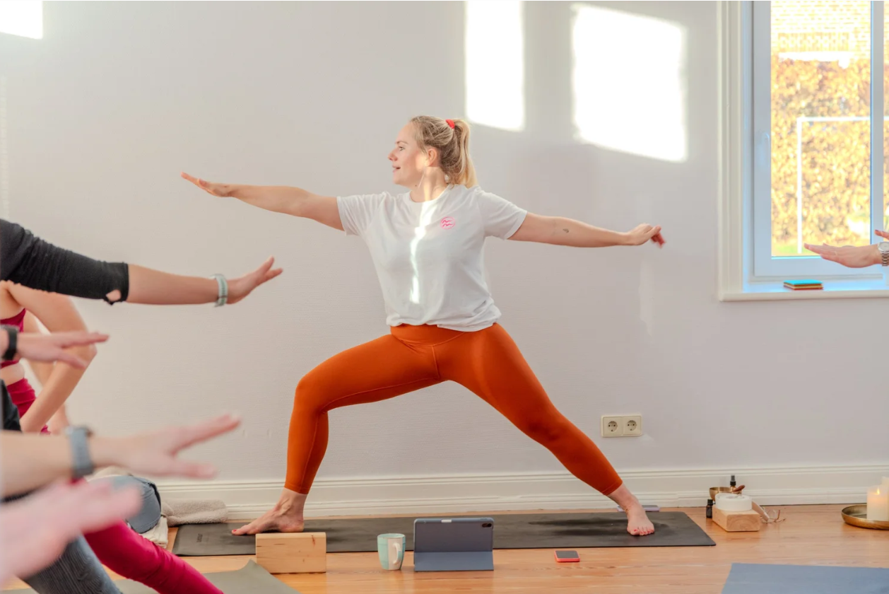Eine Frau in orangenen Leggings und weißem T-Shirt führt eine Yoga-Pose in einem hellen Raum aus. Andere Teilnehmer machen mit.