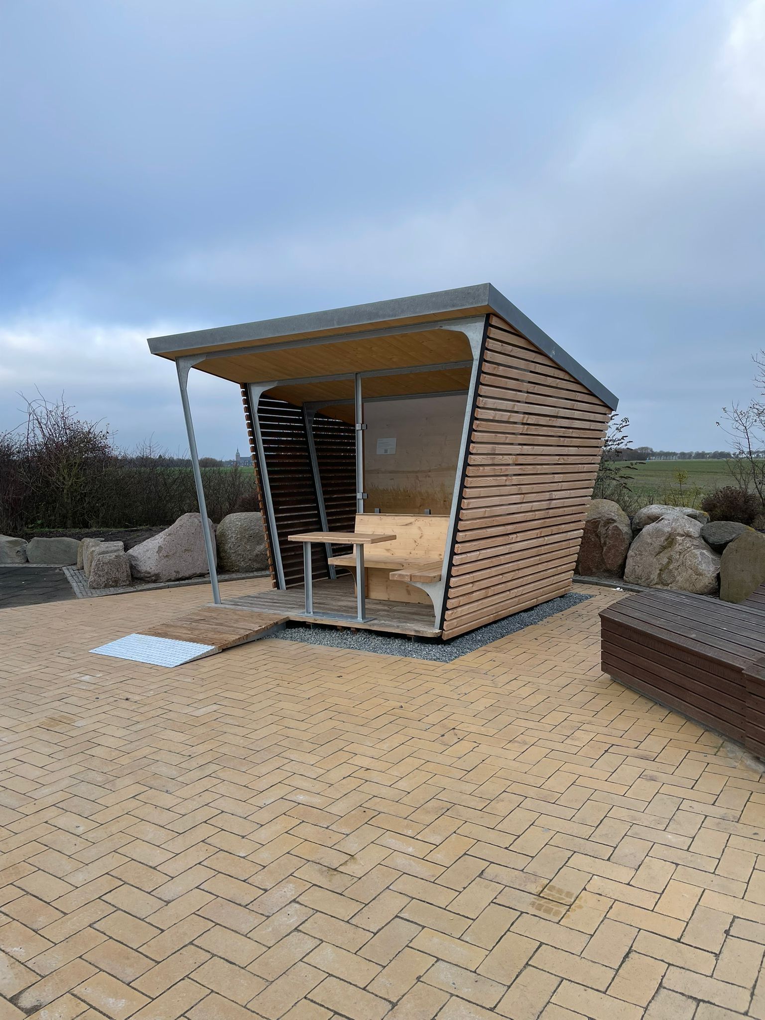 Moderne Wetterschutzhütte aus Holz mit schrägem Dach und Sitzbank auf einem gepflasterten Platz.