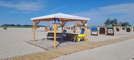 Ein barrierefreier Strandbereich mit Holzdach, Bänken und Rollstuhlzugang. Im Hintergrund sind Strandkörbe und ein blauer Himmel zu sehen.