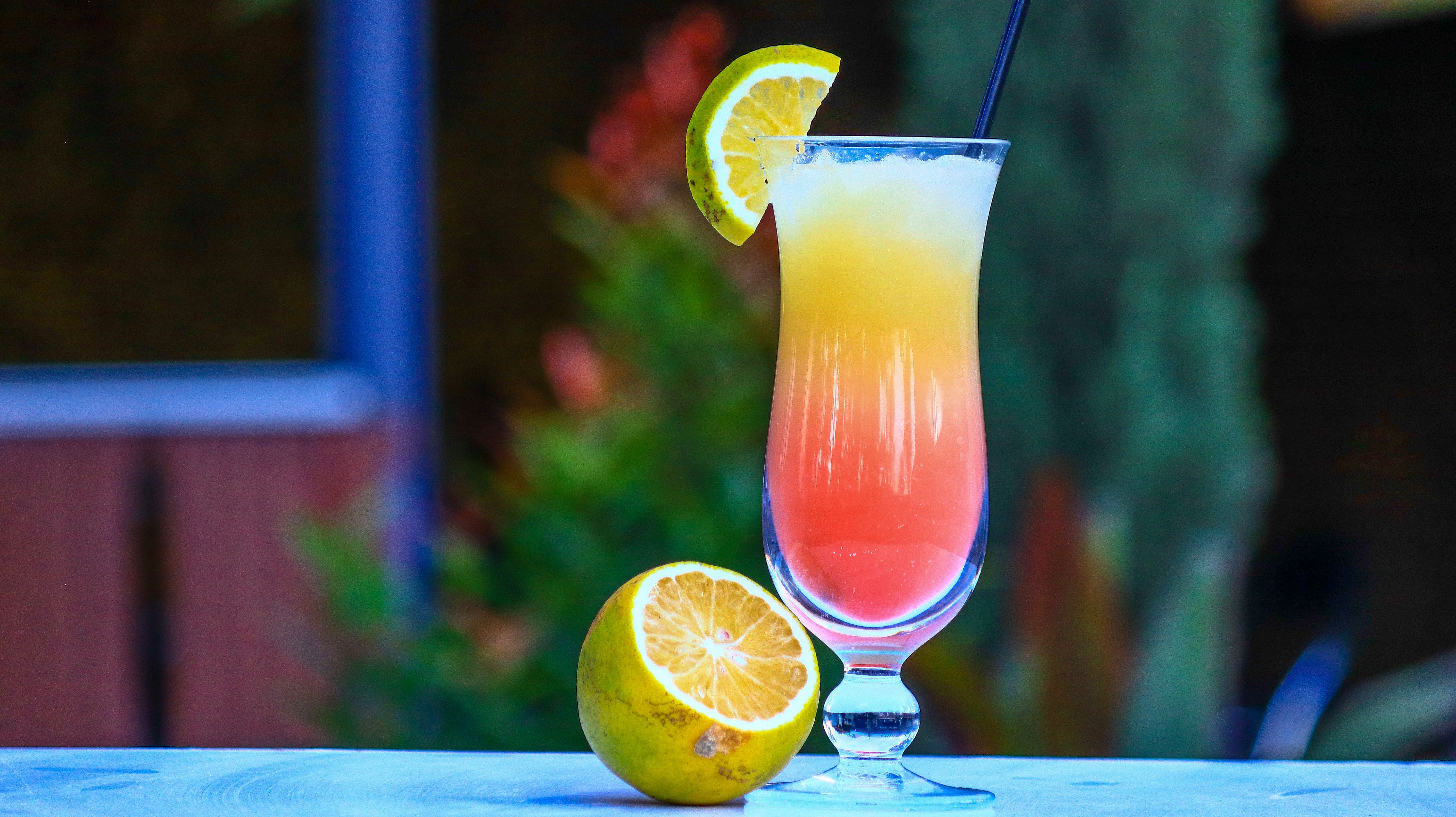 Ein Cocktailglas mit einem bunten Getränk und Zitronenscheibe, daneben eine halbe Zitrone auf einem Tisch. Der Hintergrund ist unscharf.