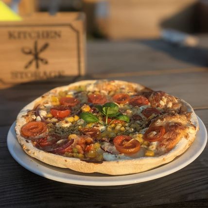 Frische Pizza mit Tomaten, Mais und Basilikum auf einem Teller, im Sonnenlicht serviert.