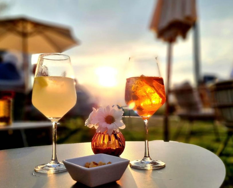 Zwei Gläser mit Getränken und eine kleine Vase mit Blumen stehen auf einem Tisch im Freien. Im Hintergrund ist ein Sonnenuntergang zu sehen.