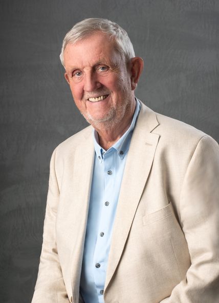 Ein älterer Mann in einem beigen Anzug und blauen Hemd lächelt freundlich vor einem grauen Hintergrund.