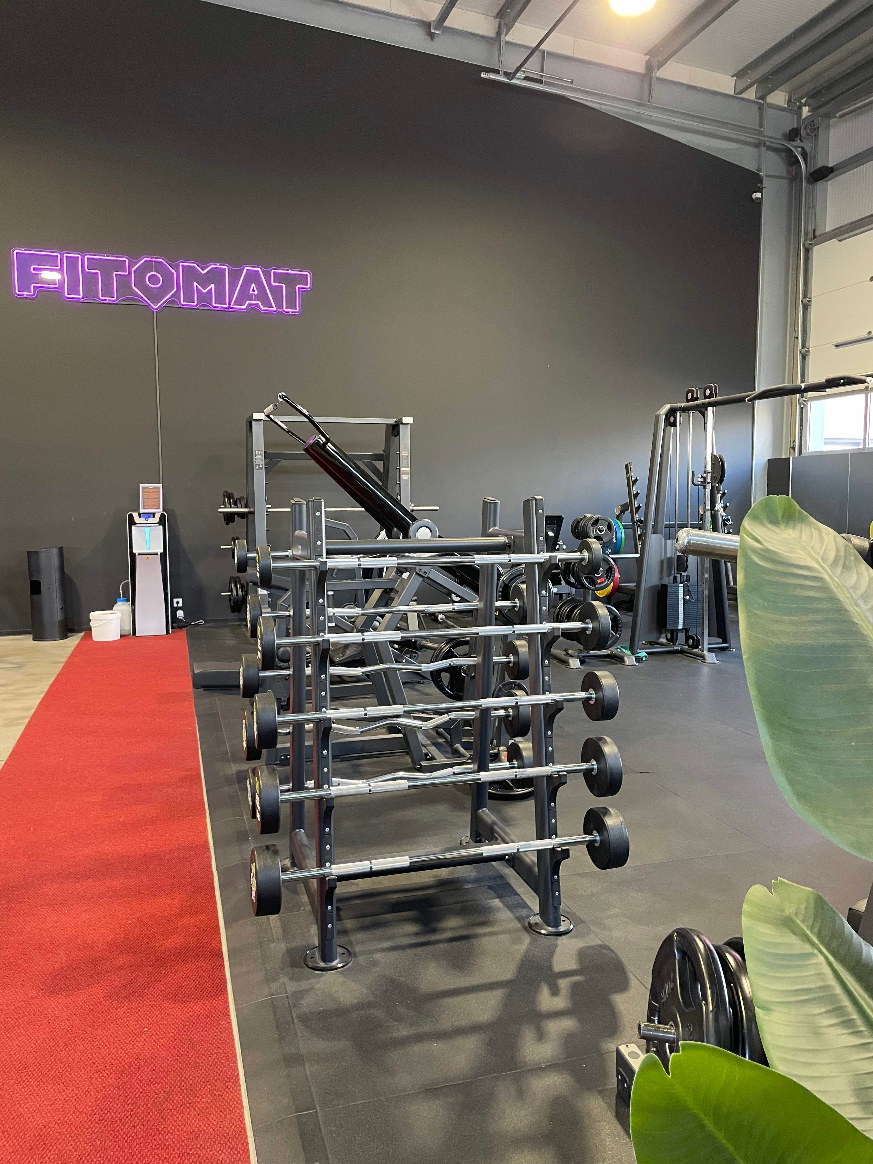 Ein Fitnessstudio mit Hanteln und Geräten, rotem Teppich und einem leuchtenden 'FITOMAT'-Schild an der Wand.