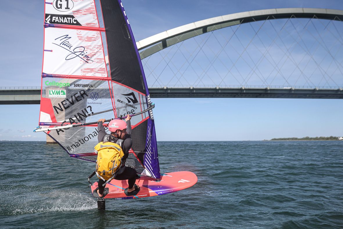 Ein Windsurfer mit gelbem Rucksack und pinkem Helm surft auf einem Foilboard vor der Fehmarnsundbrücke.