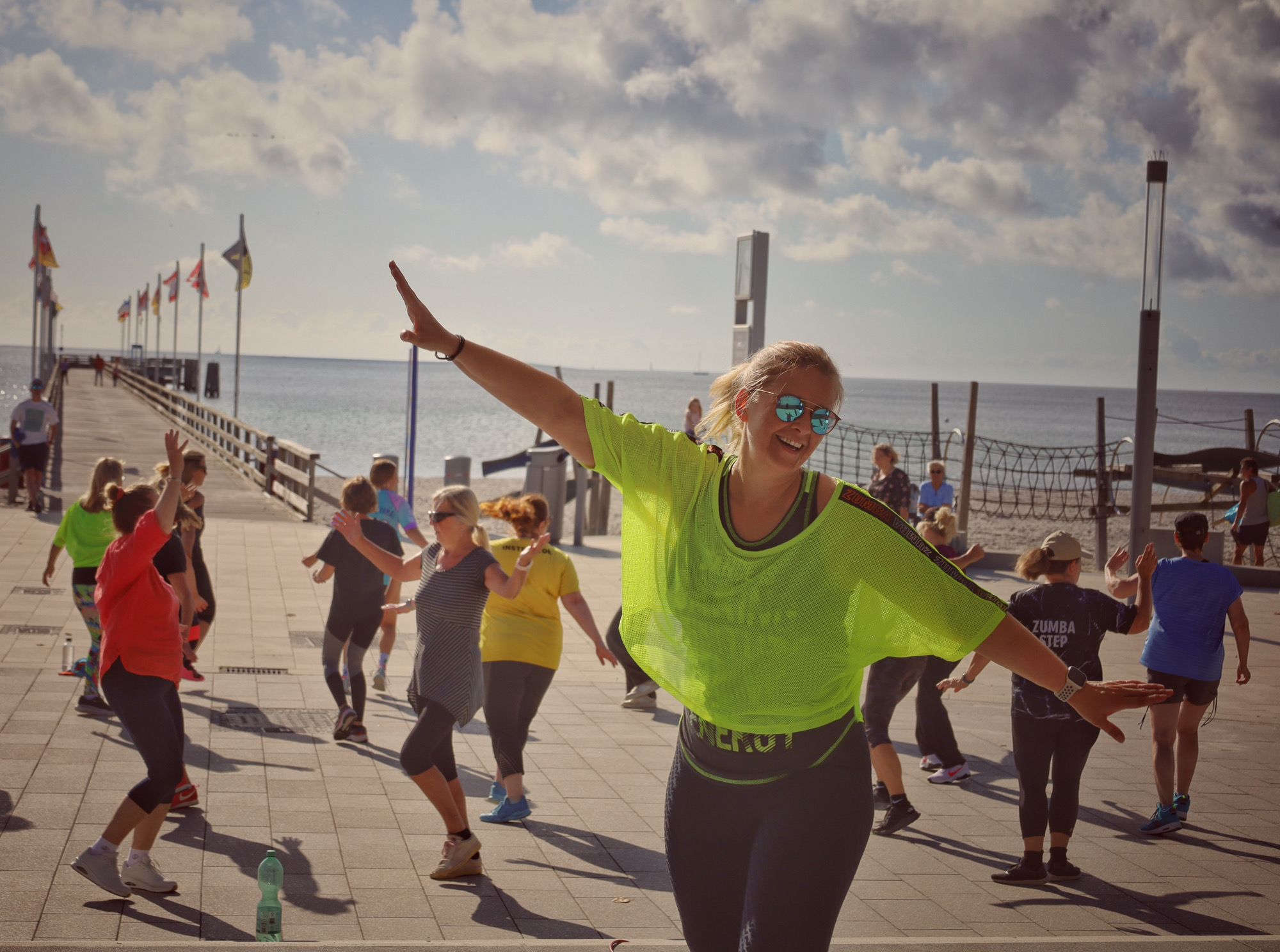 Eine Gruppe von Menschen tanzt Zumba auf einer Promenade am Meer. Im Vordergrund eine Frau in neongelbem Shirt, im Hintergrund eine Seebrücke.