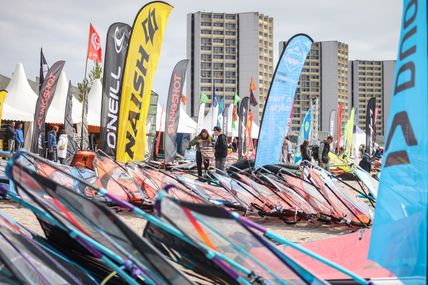 Bunte Surfsegel liegen am Strand. Im Hintergrund wehen bunte Fahnen