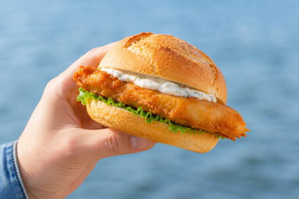 Ein knuspriges Fischbrötchen mit Salat und Sauce wird vor einem blauen Gewässer in der Hand gehalten.