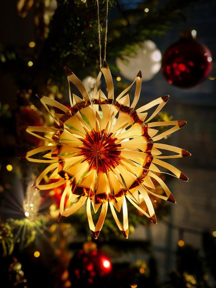 Ein kunstvoll gefertigter Strohstern hängt an einem Weihnachtsbaum, umgeben von roten und weißen Kugeln. Warmes Licht erzeugt eine festliche Atmosphäre.