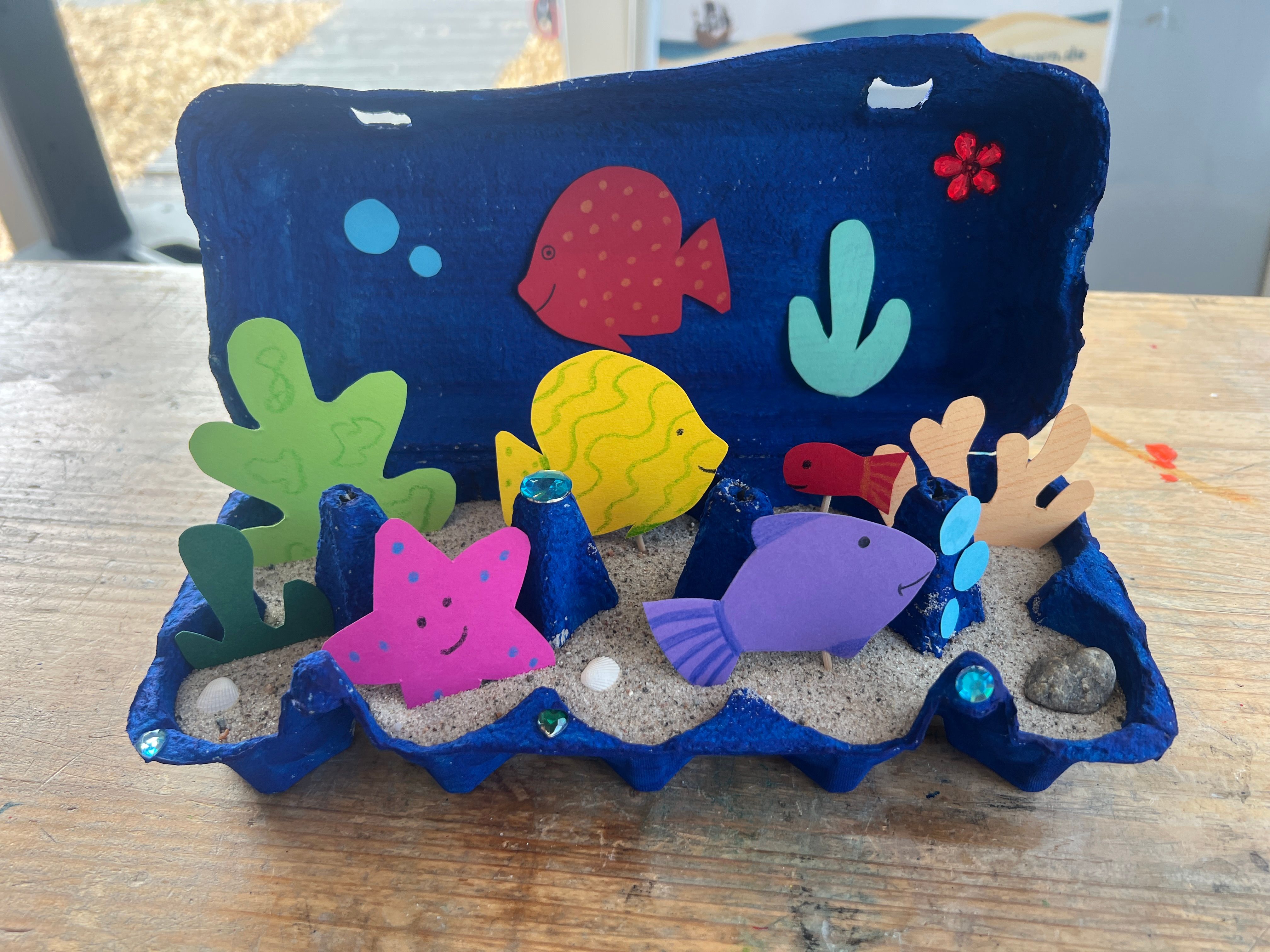 Ein kreatives Aquarium aus einem blauen Eierkarton mit bunten Papierfischen und Pflanzen, dekoriert mit Sand und kleinen Steinen.