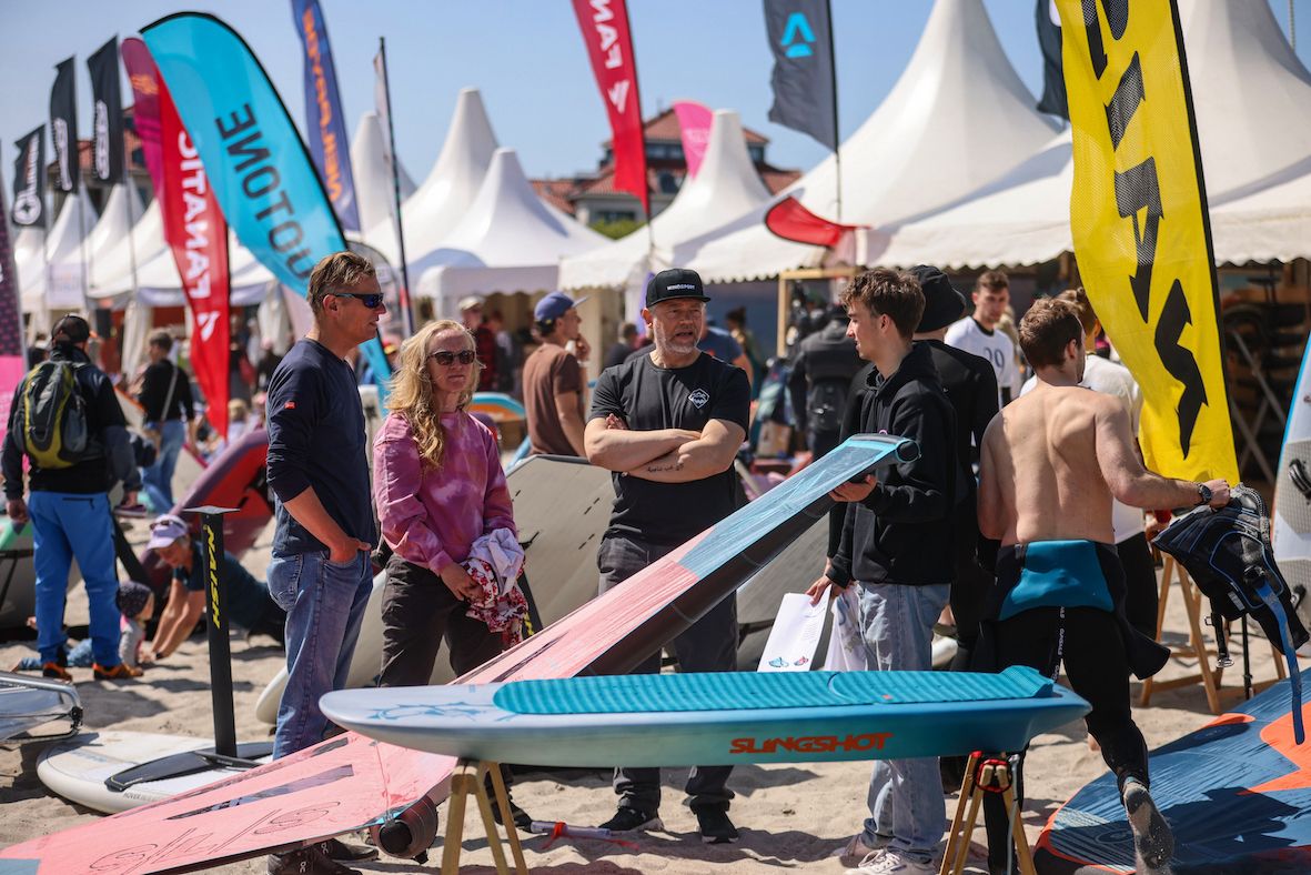 Menschen auf einem Surf-Festival mit Surfbrettern, Zelten und bunten Fahnen im Hintergrund. Einige Personen unterhalten sich, andere betrachten die Ausrüstung.