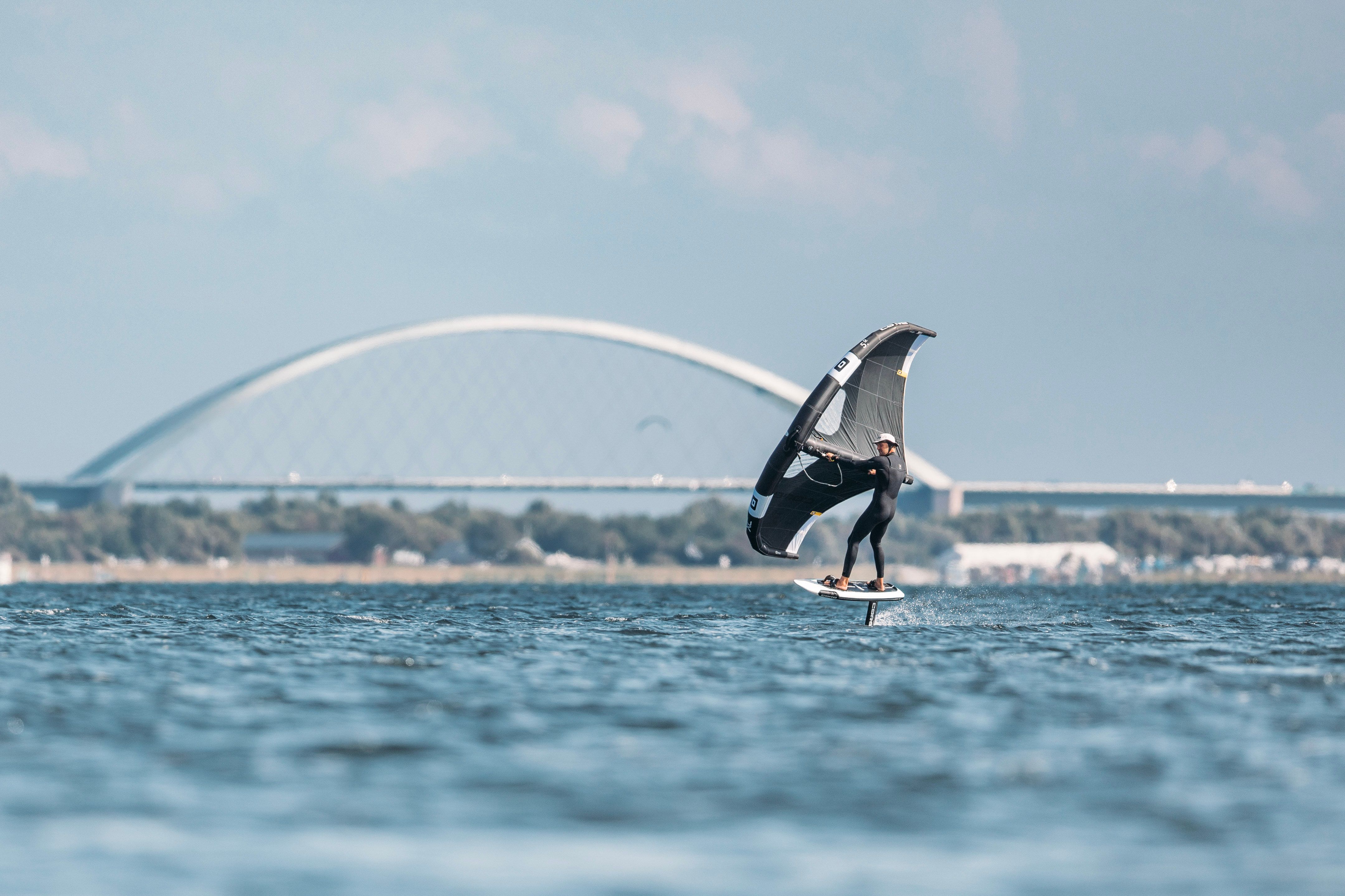 Ein Wingfoiler gleitet über das Wasser vor der Fehmarnsundbrücke. Der Himmel ist klar und das Wasser ruhig.