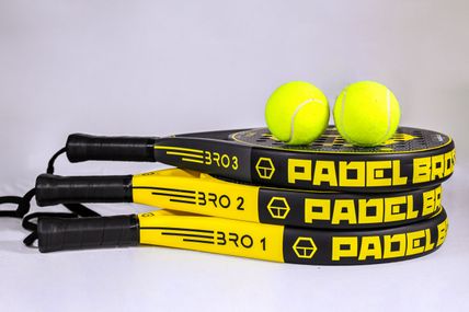 Drei gelb-schwarze Padel-Schläger liegen gestapelt, mit zwei gelben Bällen darauf. Die Schläger sind mit 'PADEL BRO' beschriftet.