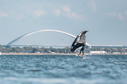 Ein Wingfoiler gleitet über das Wasser vor der Fehmarnsundbrücke. Der Himmel ist klar und das Wasser ruhig.