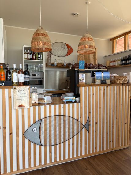 Ein gemütliches Café mit Holztresen, dekoriert mit Fischmotiven. Hängelampen aus Korbgeflecht und eine Auswahl an Getränken sind sichtbar.
