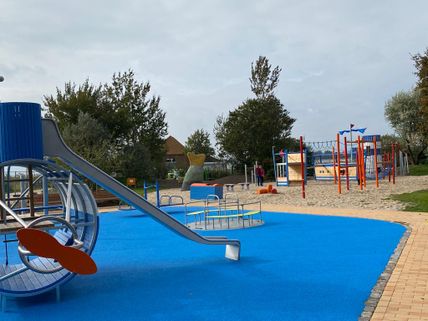 Ein moderner Spielplatz mit blauen und roten Spielgeräten, darunter eine Rutsche und Klettergerüste, auf einem blauen Bodenbelag. Im Hintergrund sind Bäume zu sehen.