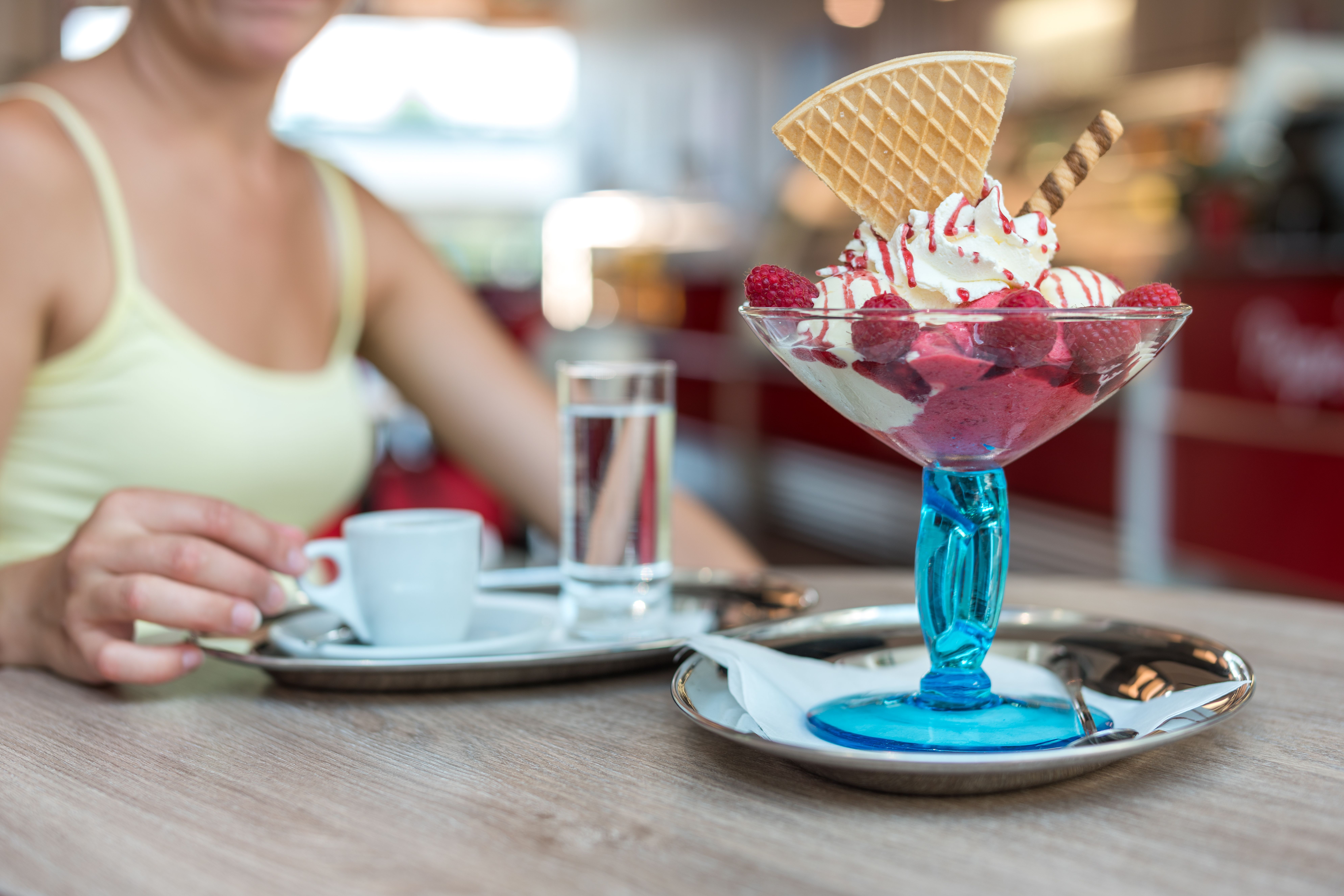 Ein köstliches Dessert mit Eiscreme, Himbeeren und Waffeln in einem blauen Glas. Im Hintergrund genießt eine Person eine Tasse Kaffee.