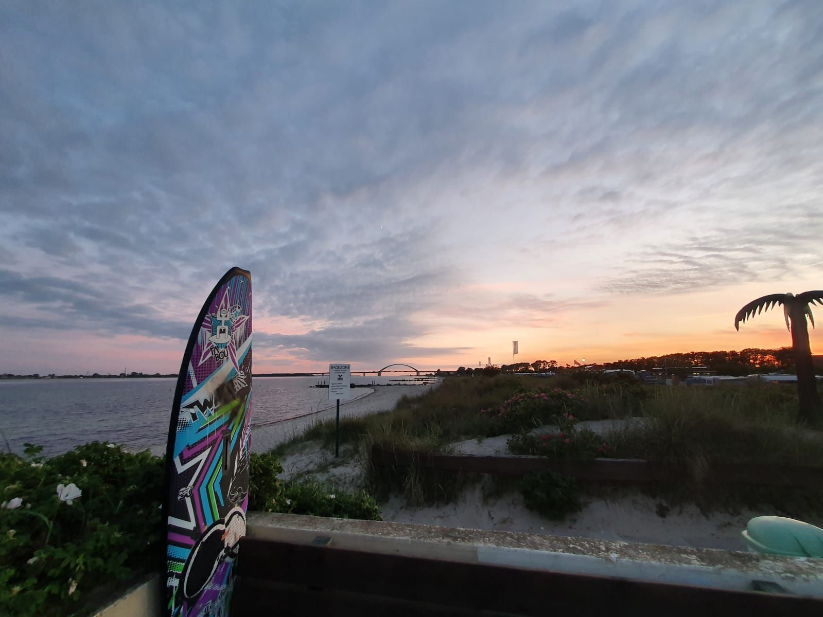 Ein farbenfrohes Surfbrett steht am Strand bei Sonnenuntergang. Eine künstliche Palme und eine Brücke sind im Hintergrund zu sehen.