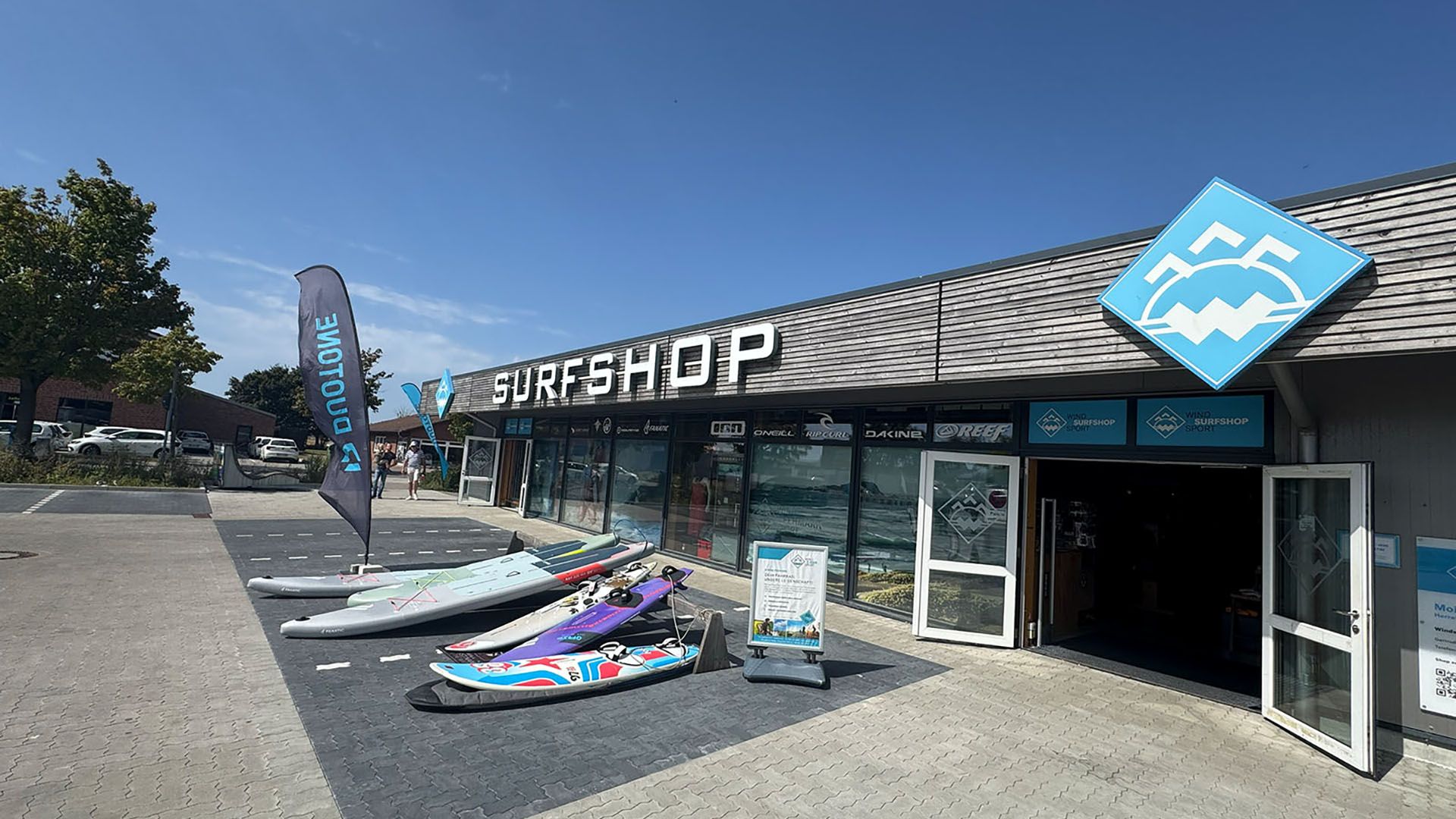 Ein Surfshop auf Fehmarn mit Surfbrettern und Flaggen vor dem Eingang. Das Gebäude hat ein modernes Design mit großen Fenstern.