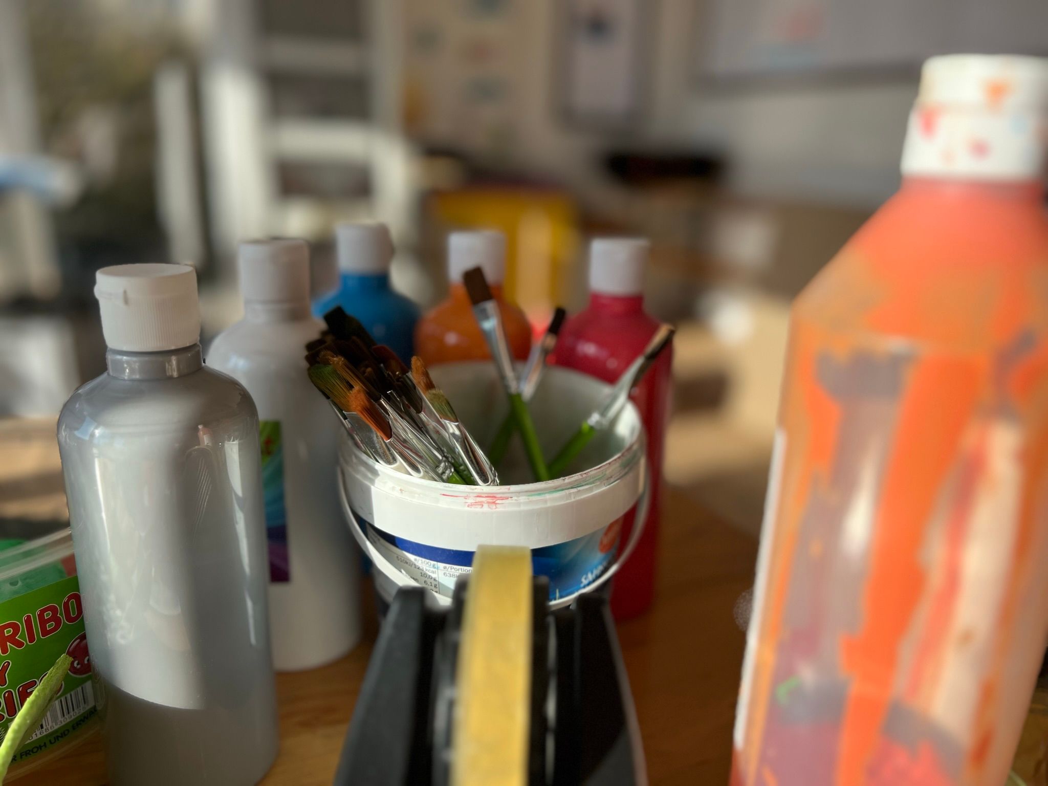 Malutensilien auf einem Tisch: Pinsel in einem Becher, umgeben von bunten Farbtuben. Sonnenlicht fällt auf die Szene und erzeugt Schatten.