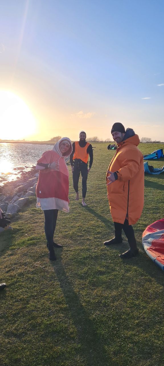 Drei Personen in Kitesurf-Ausrüstung stehen auf einer Wiese am Wasser. Der Sonnenuntergang im Hintergrund taucht die Szene in warmes Licht.