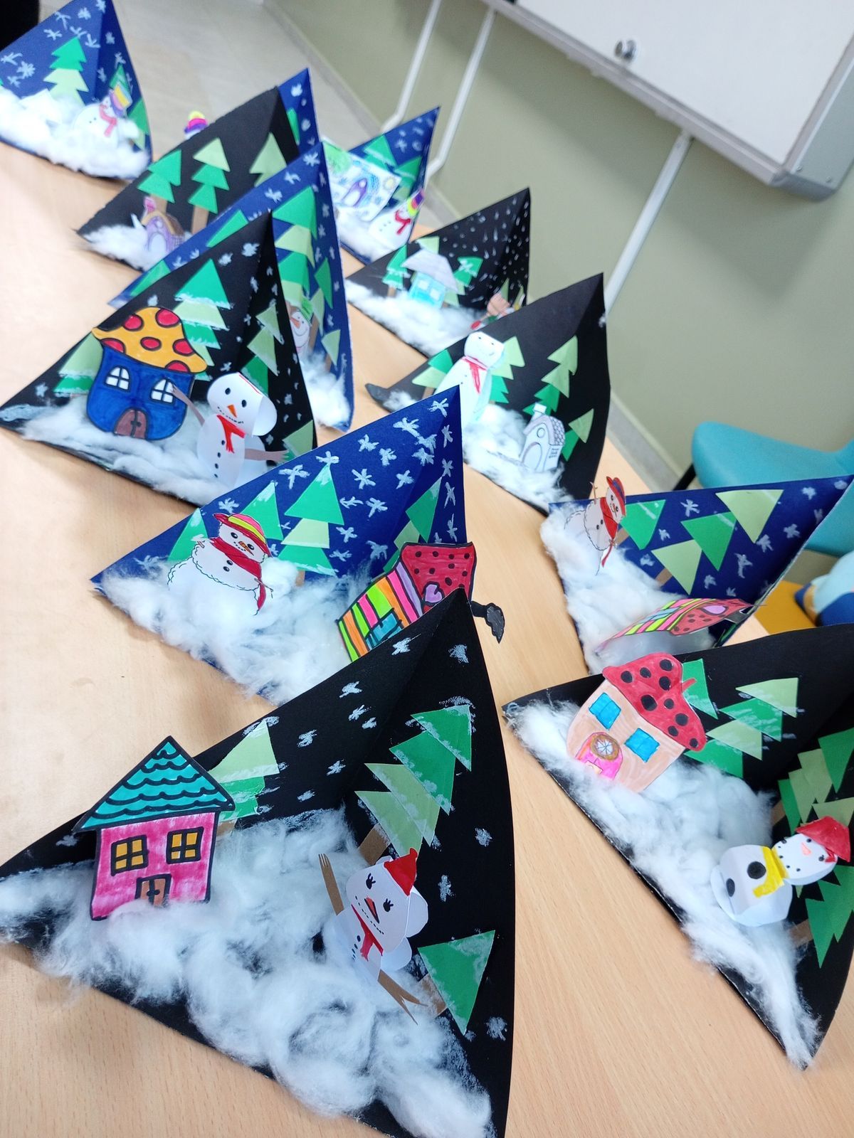 Bunte Bastelarbeiten zeigen ein Winterdorf mit Schneemännern und Häusern aus Papier, dekoriert mit Watte als Schnee, auf einem Tisch.