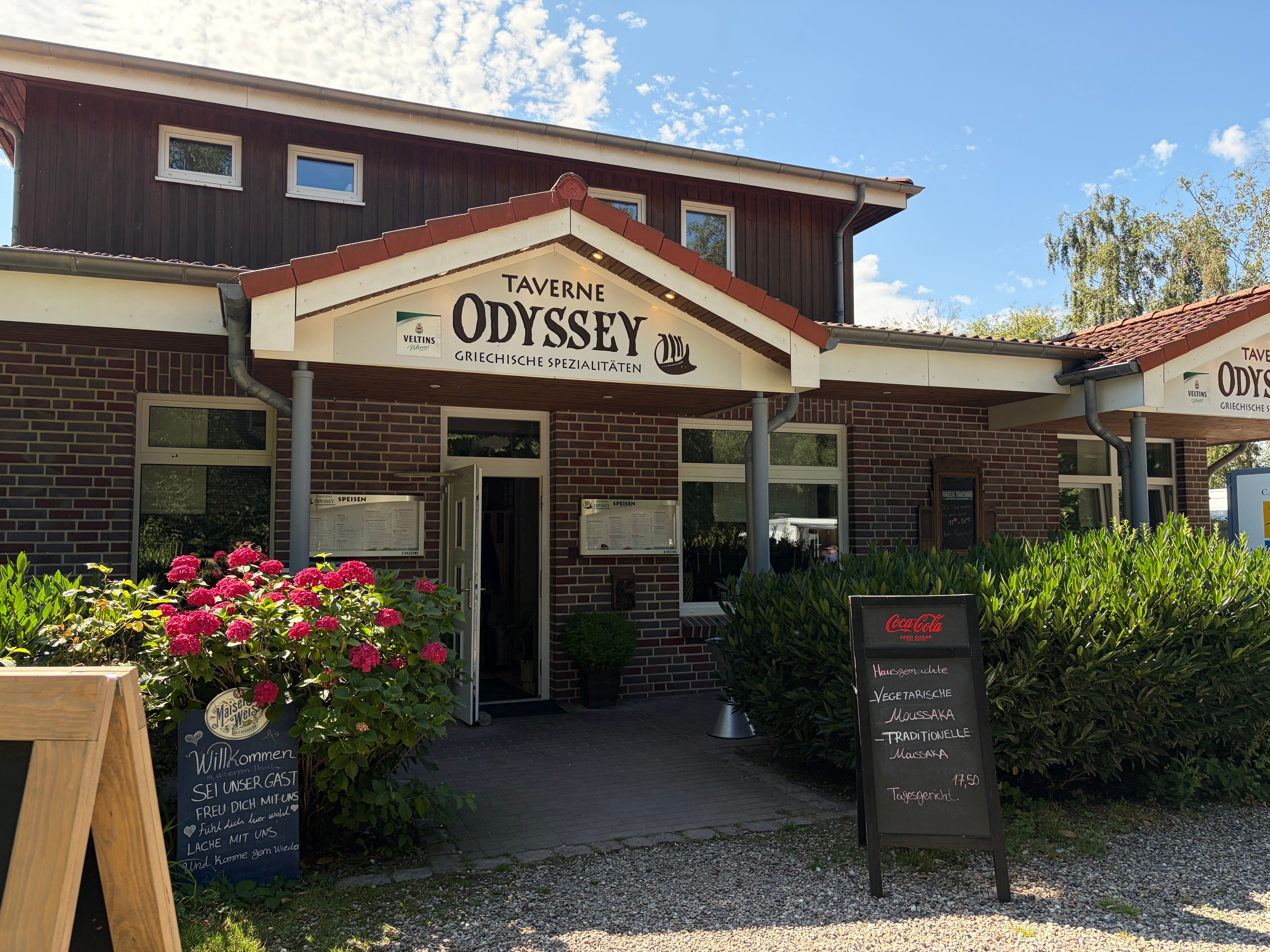 Die Taverne Odyssey bietet griechische Spezialitäten an. Vor dem Eingang stehen Schilder mit Tagesgerichten und ein Blumenbeet.