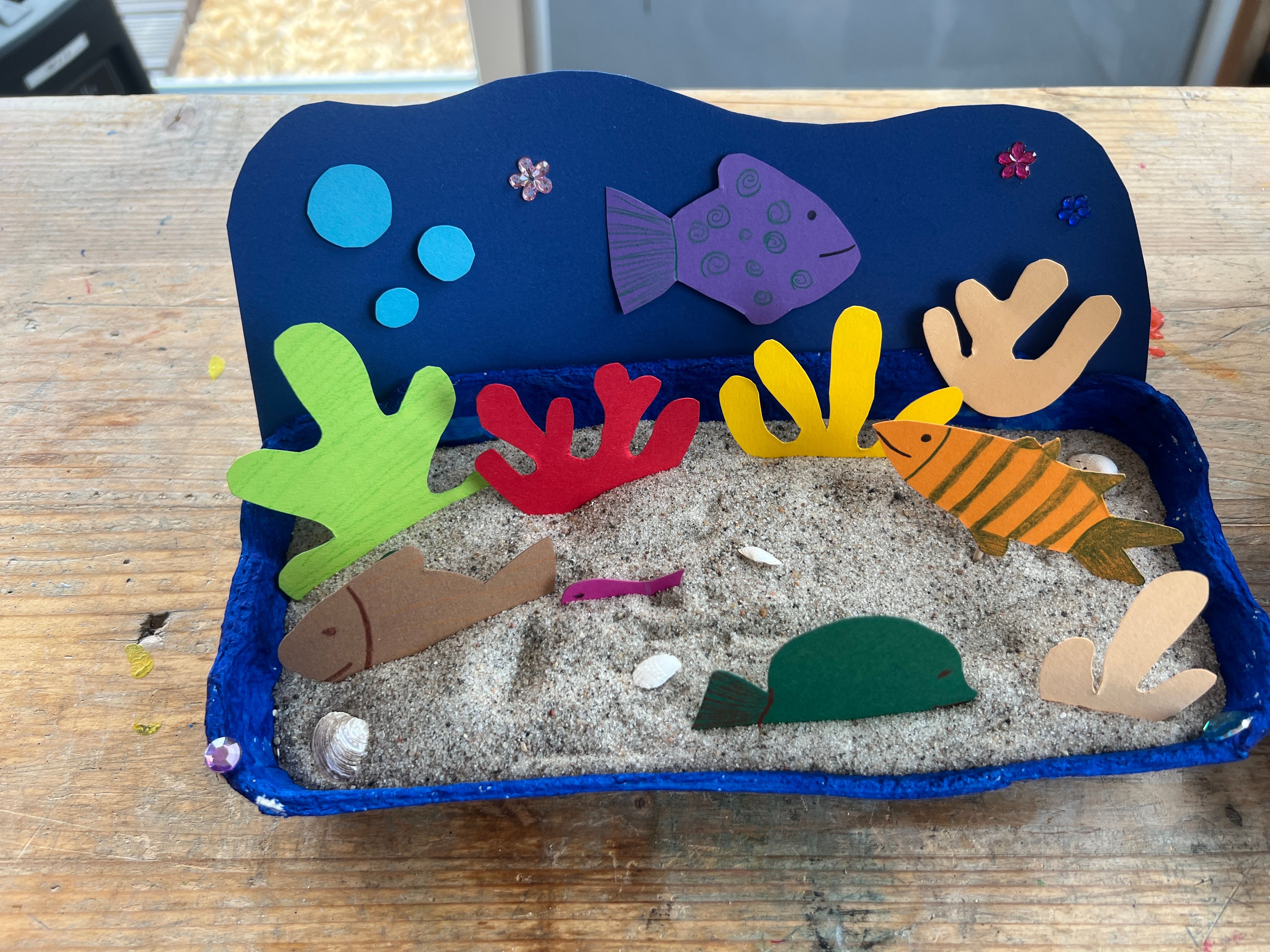 Ein kreatives DIY-Aquarium aus Papier mit bunten Fischen und Korallen auf Sand in einer blauen Schale.