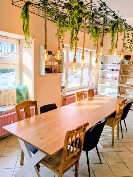 Ein gemütlicher Raum mit einem langen Holztisch und verschiedenen Stühlen. Über dem Tisch hängen Glühbirnen und Pflanzen, die eine warme Atmosphäre schaffen.