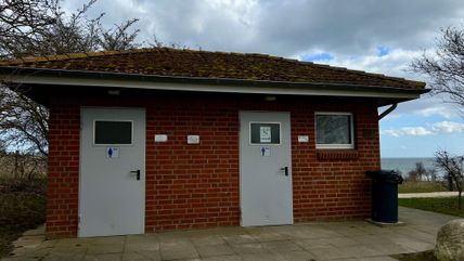 Backsteingebäude mit zwei Toilettentüren, umgeben von Bäumen und mit Blick auf das Meer. Der Himmel ist bewölkt.