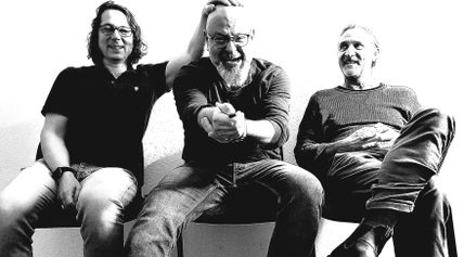 Drei Männer sitzen nebeneinander und lachen. Der mittlere Mann wird von einem der anderen am Kopf berührt. Schwarz-Weiß-Foto mit hohem Kontrast.