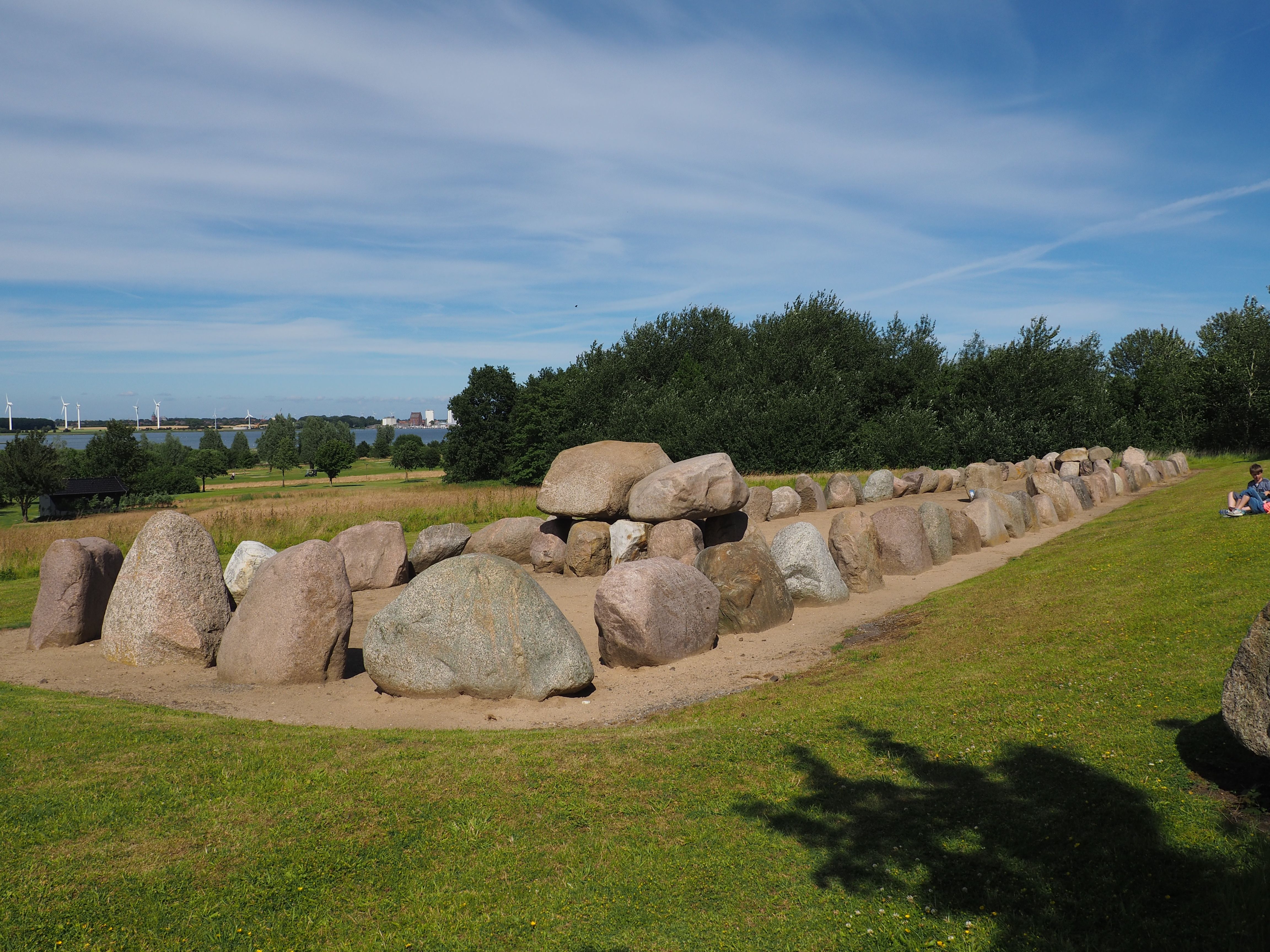 Eine lange Reihe großer Steine auf einer grünen Wiese, umgeben von Bäumen. Im Hintergrund sind Windräder und ein klarer Himmel zu sehen.