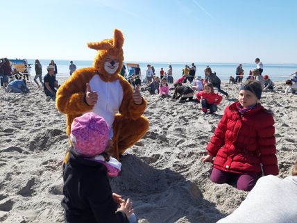 Ein Mensch im Hasenkostüm gibt Daumen hoch am Strand, umgeben von spielenden Kindern im Sand. Der Himmel ist klar und blau.