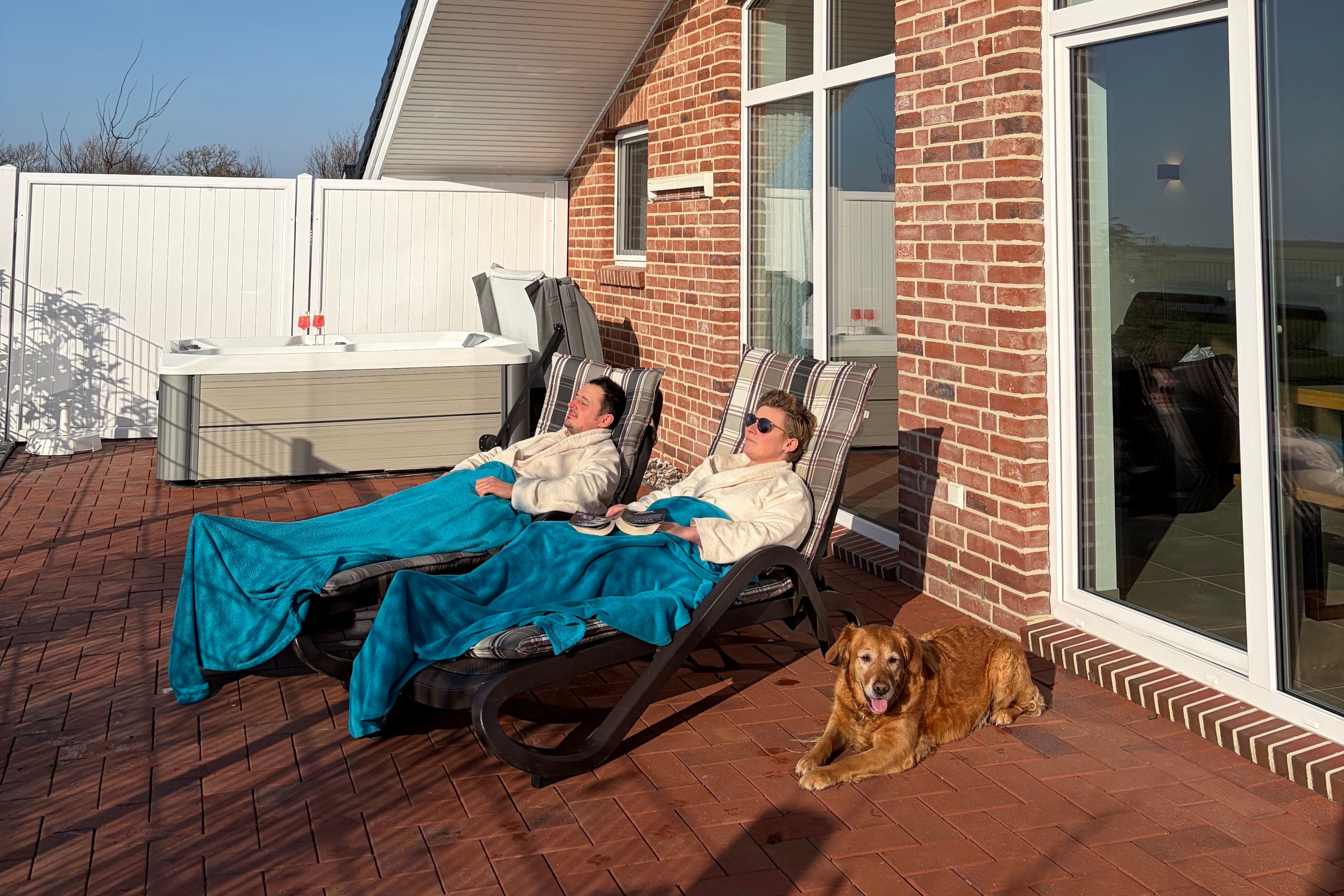 Zwei Personen liegen auf Liegestühlen auf einer Terrasse neben einem Hund und einem Whirlpool vor einem Backsteinhaus.