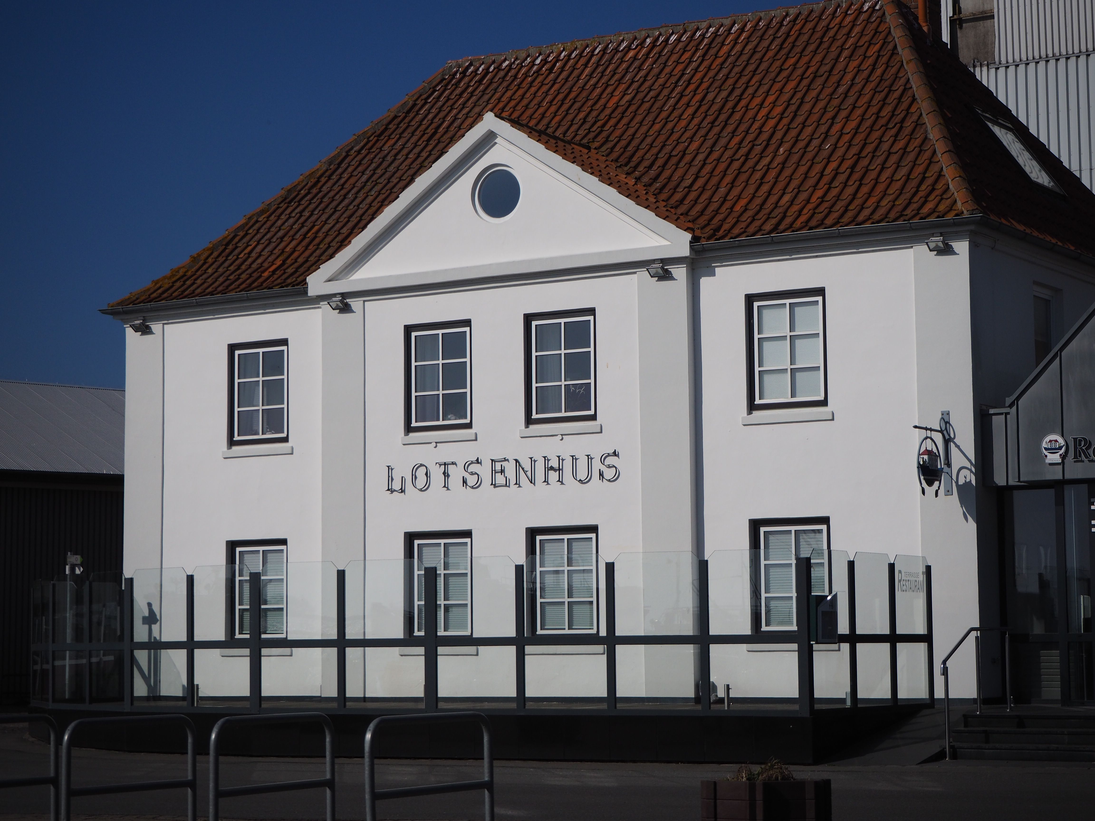 Das Lotsenhus in Burgstaaken ist ein weißes Gebäude mit rotem Ziegeldach und der Aufschrift 'Lotsenhus'. Es steht unter klarem, blauem Himmel.