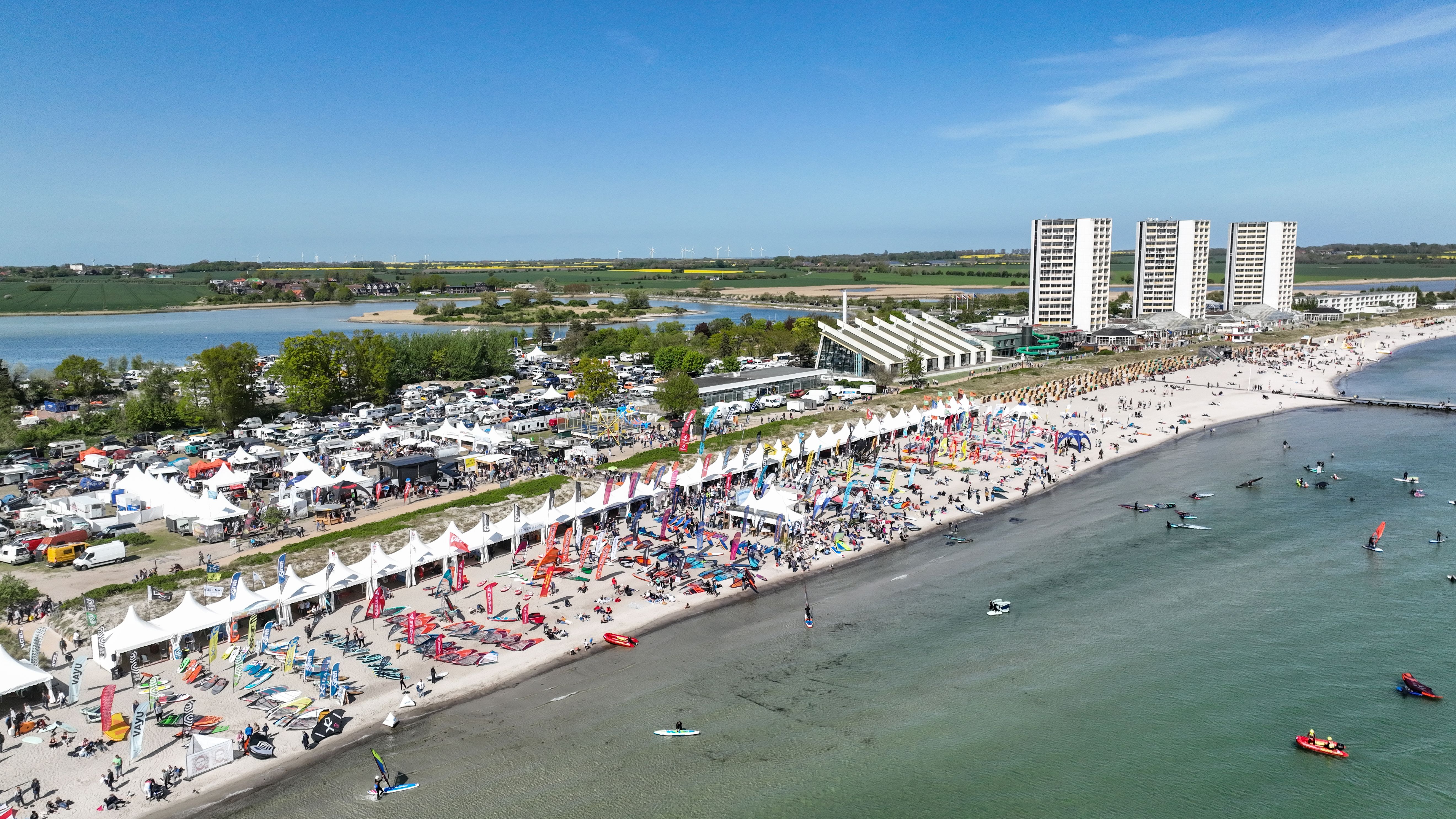 Luftaufnahme eines Strandes mit Surffestival. Viele bunte Zelte und Surfbretter am Strand, Menschen im Wasser. Im Hintergrund hohe Gebäude und grüne Landschaft.