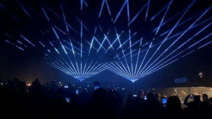 Eine Menschenmenge beobachtet eine beeindruckende Lasershow mit blauen Strahlen in der Nacht. Viele halten ihre Handys hoch.